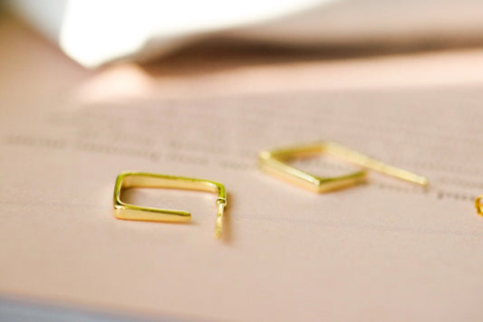 A pair of Sterling Silver/Gold Vermeil geometric square hoop earrings with a minimalist modern design, resting on a soft pink and white background.